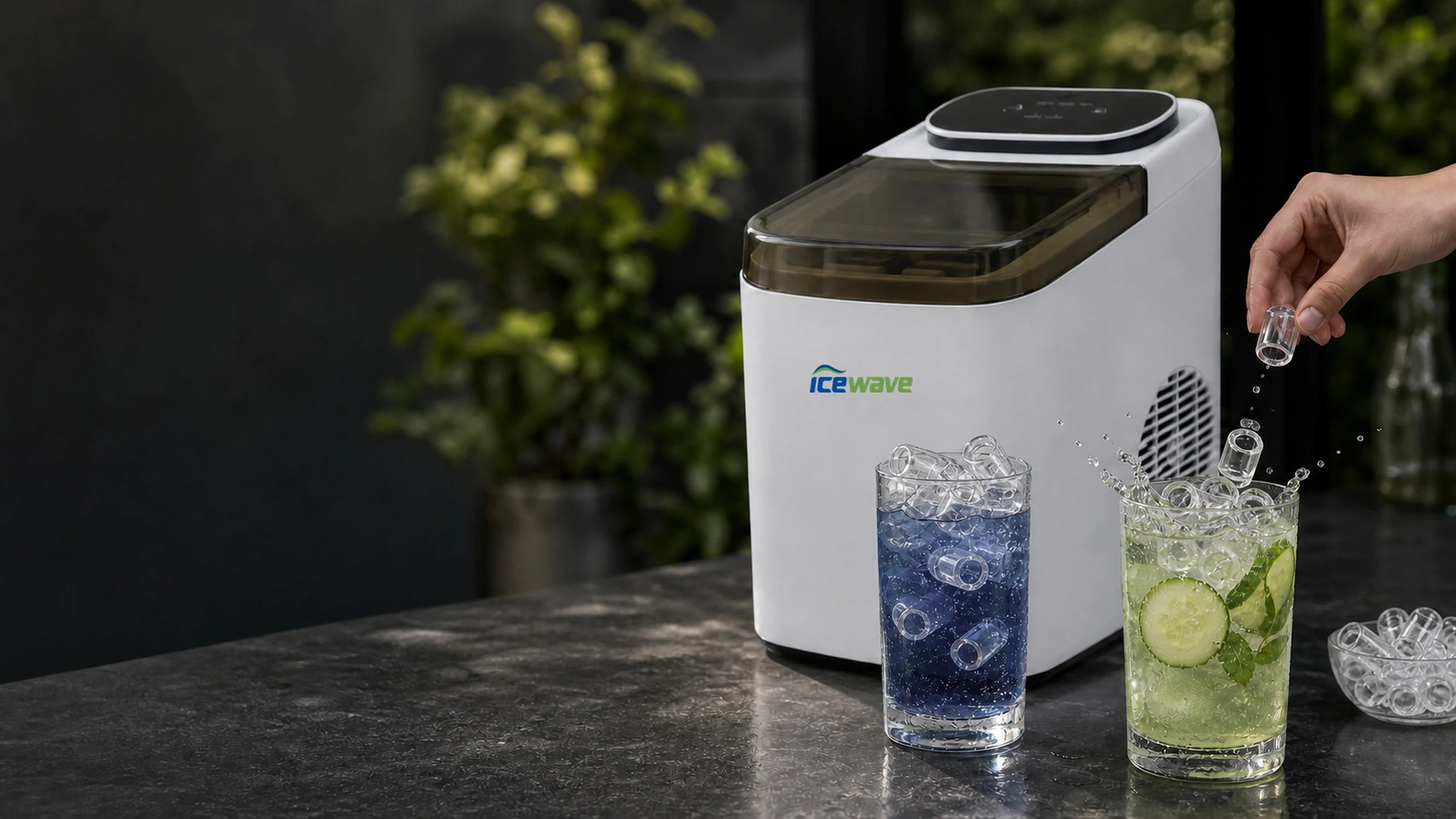 ZB-13 countertop ice maker on dark stone counter, hand adding bullet ice to a tall glass of sparkling water with cucumber, lime and fresh mint, paired with a blueberry sparkling drink on the side — modern home bar with green plants blurred in background.
